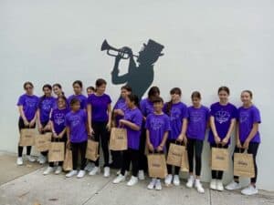 Young girls in purple GEM shirts holding WDNA bags, standing by a jazz mural, symbolizing mentorship and opportunity in music and media.
